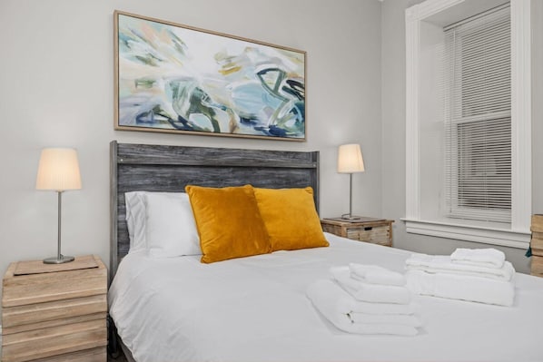 A neatly made bed with white bedding and a mustard-colored pillow, flanked by two bedside tables with lamps, in a room with a large abstract painting above the bed and a window with blinds to the right. - Walk to Forest Park Fire Pit Porch Swing (St. Louis)