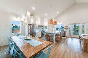 A modern kitchen with a central island, stainless steel appliances, and a dining area with a wooden table and blue chairs. - Glory Daze Private Pool Ice Maker EV Charger (Port Aransas)