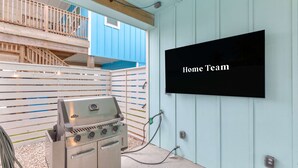 A barbecue grill with a TV screen displaying 'Home Team' in a covered outdoor area with a wooden deck and a blue wall. - Glory Daze Private Pool Ice Maker EV Charger (Port Aransas)