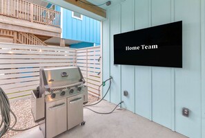 Ein Grill mit einem Fernsehbildschirm, der 'Home Team' zeigt, in einem überdachten Außenbereich mit Holzterrasse und blauer Wand.