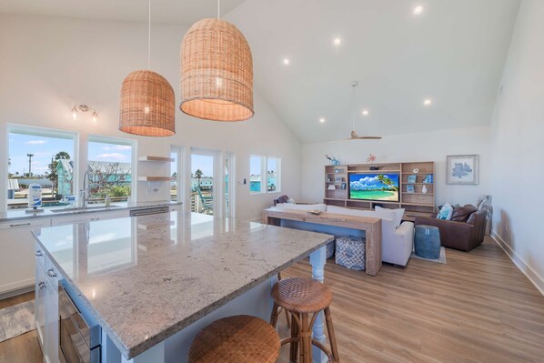 A spacious living area with a large island, wicker pendant lights, a flat-screen TV, and a view of the outdoors. - Glory Daze Private Pool Ice Maker EV Charger (Port Aransas)