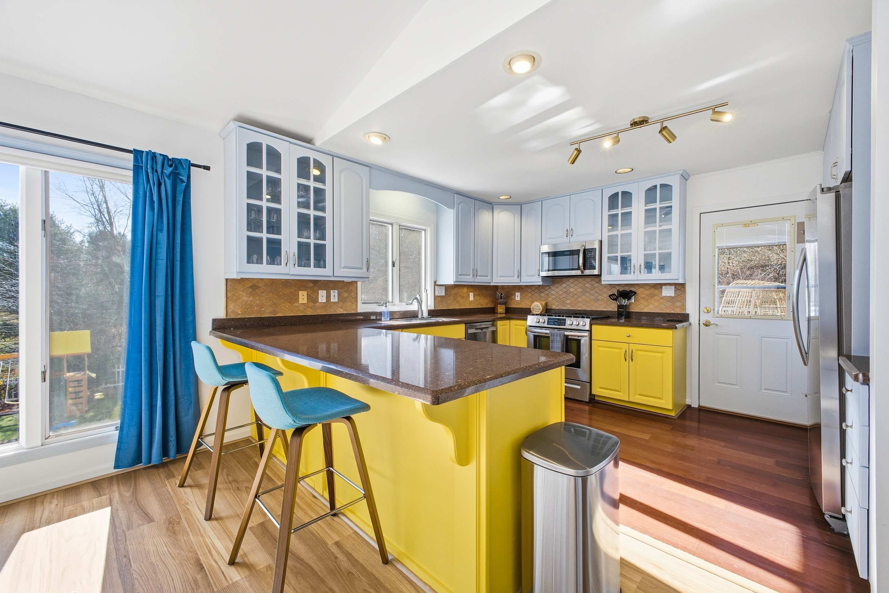 A modern kitchen with a central island, bar stools, a stainless steel refrigerator, and a large window with curtains.