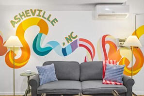 A room with a grey sofa, two lamps, a wall with colorful abstract art, and an air conditioning unit. - Sleeps 20 Sport Court Game Room Sauna Spa (Asheville)