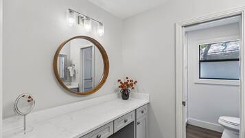 A bathroom with a white vanity, a round mirror with a wooden frame, a vase with flowers, and a window with a view of trees.