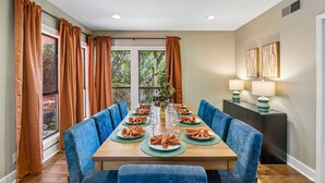 A dining room with a long table, blue upholstered chairs, and a view of trees through large windows. - The Haven on Twisted Wood 4BR 3BA Sleeps 12 (San Antonio)