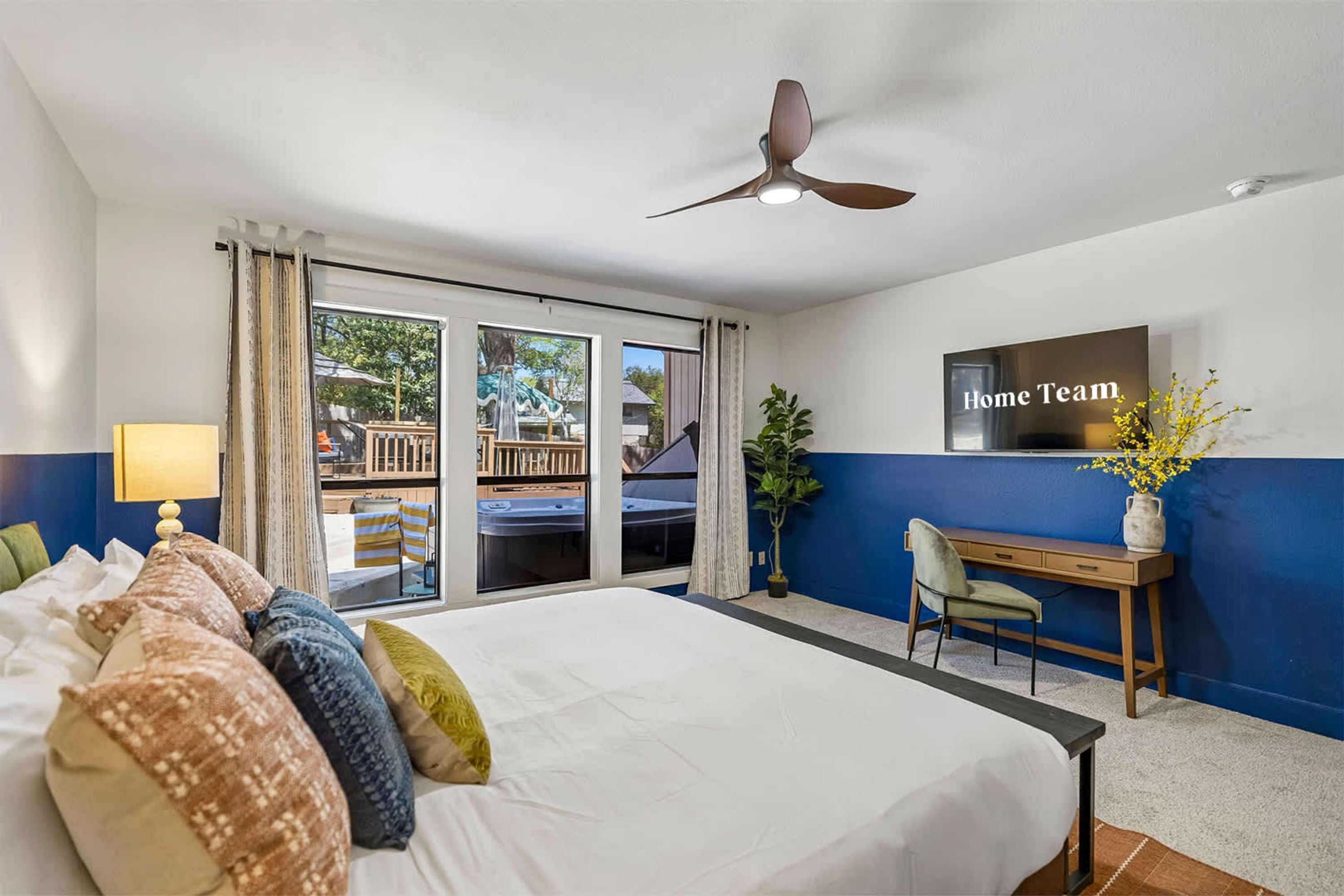 A hotel room with a bed, a desk, a chair, a ceiling fan, and a view of the outside.