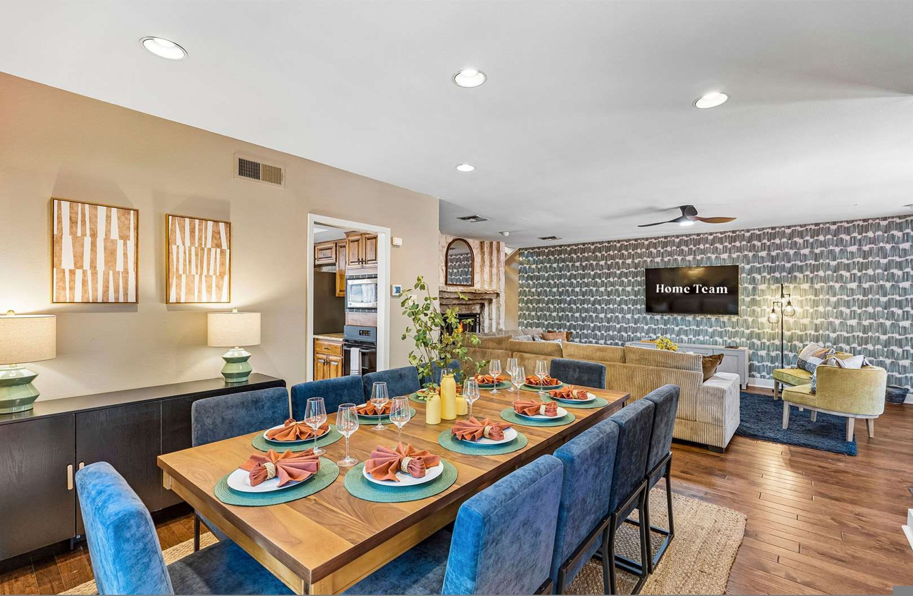 A modern dining area with a long table set for six, blue upholstered chairs, and a kitchen visible in the background.