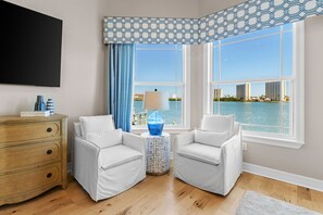 A room with two white armchairs, a wooden dresser, a round side table with a blue vase, and a view of the water and buildings through large windows. - Luxury Waterfront 4Br Private Heated Pool Hot Tub (St. Petersburg)
