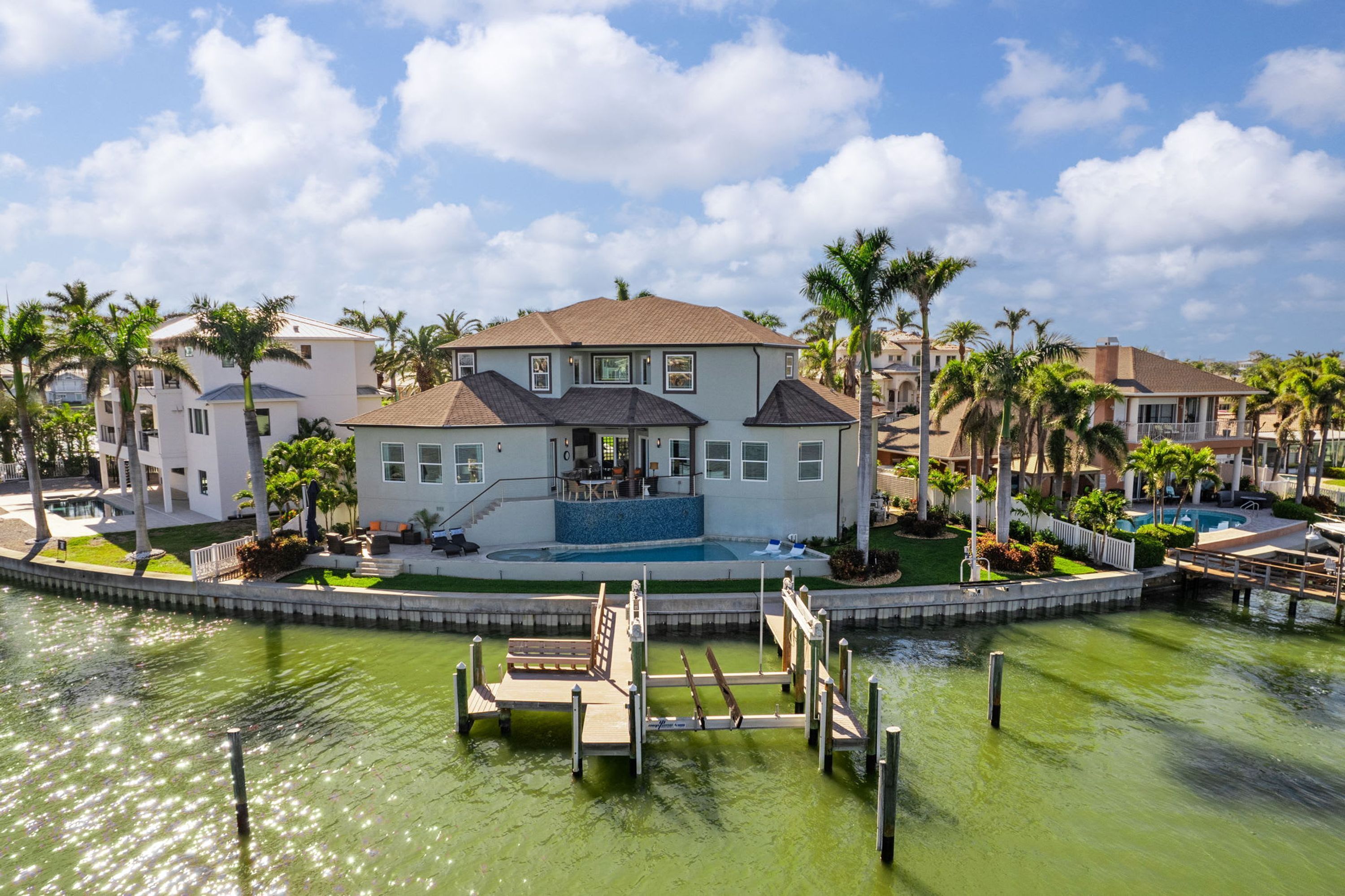 A two-story house with a swimming pool, surrounded by palm trees and a dock.