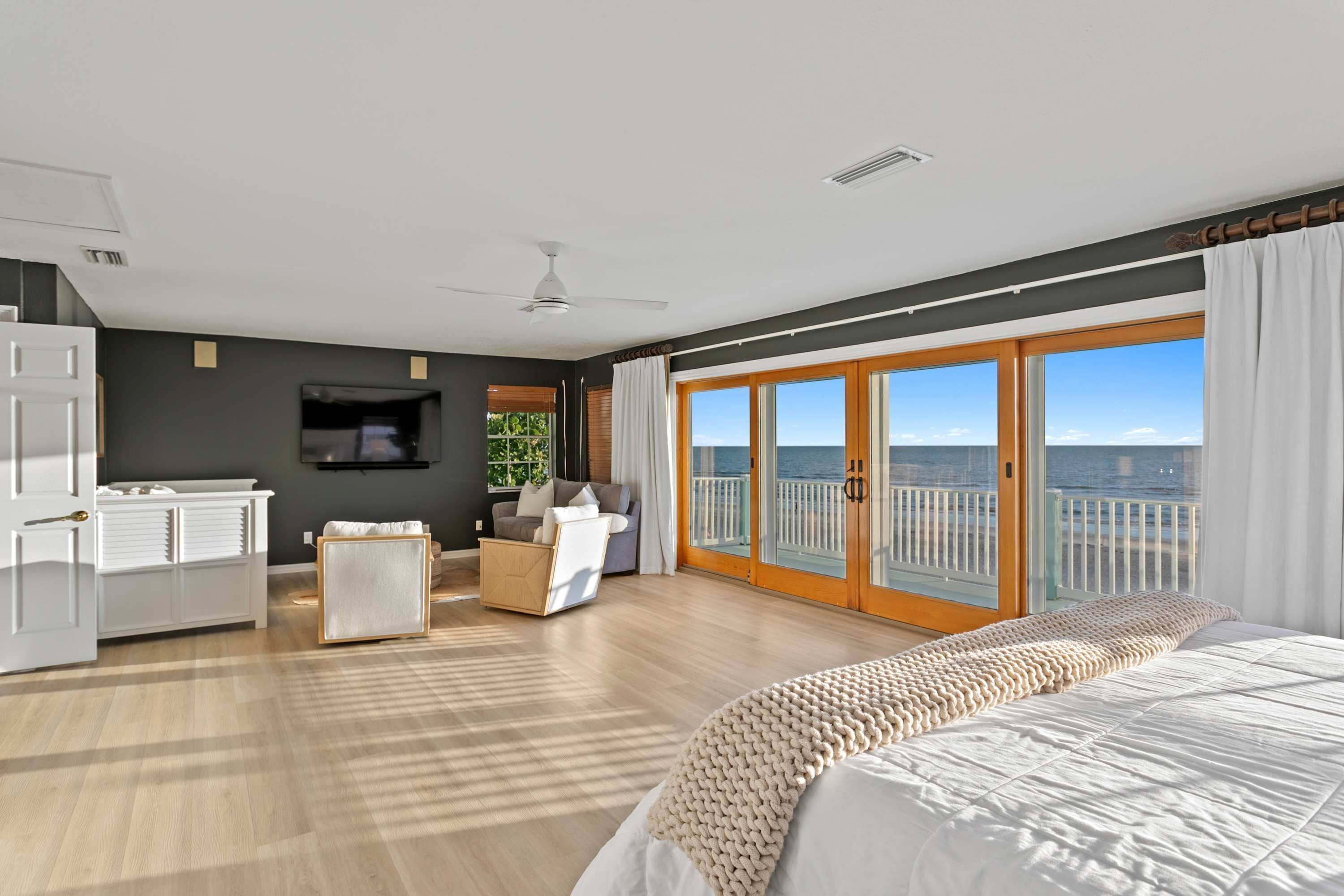 A hotel room with a bed, a seating area with a sofa and armchairs, a television, and a balcony with a view of the sea.