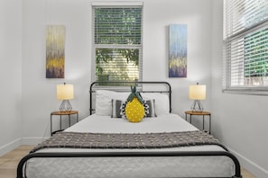 Ein Schlafzimmer mit einem Bett, zwei Nachttischlampen, einem Fenster mit Jalousien und zwei abstrakten Gemälden an der Wand.