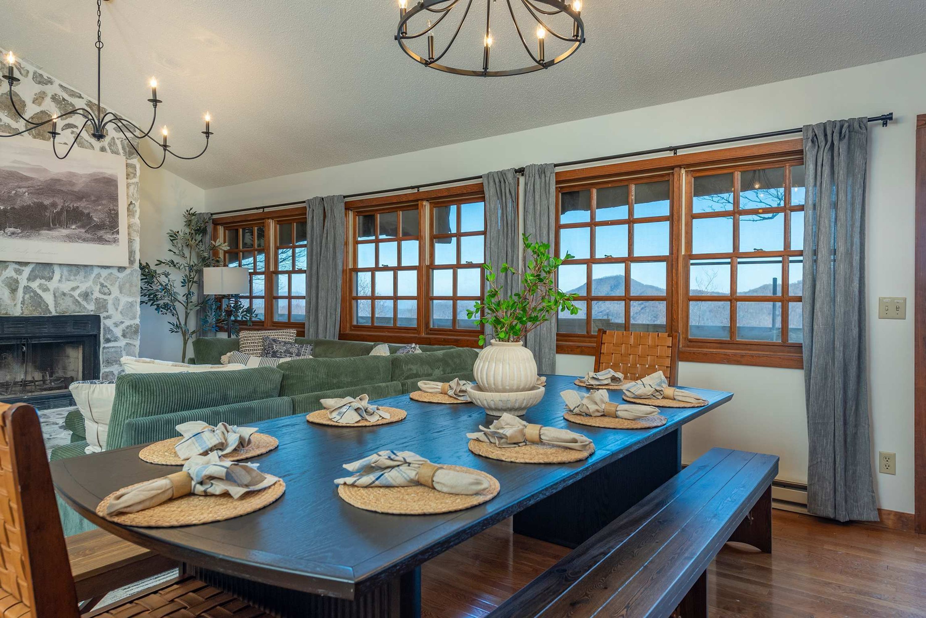 A dining area with a large table set for four, flanked by windows with curtains, a stone fireplace, and a hanging light fixture.