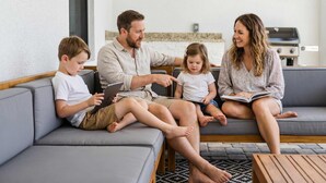 A family of four seated on a grey sofa in a modern living room with a kitchen in the background. - Backyard Oasis Pool Game Room Sleeps 10 (Cape Coral)