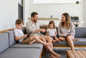 A family of four seated on a grey sofa in a modern living room with a kitchen in the background. - Backyard Oasis Pool Game Room Sleeps 10 (Cape Coral)