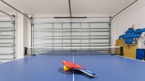 A ping pong table with a net in a garage-like space with metal garage doors. - Backyard Oasis Pool Game Room Sleeps 10 (Cape Coral)