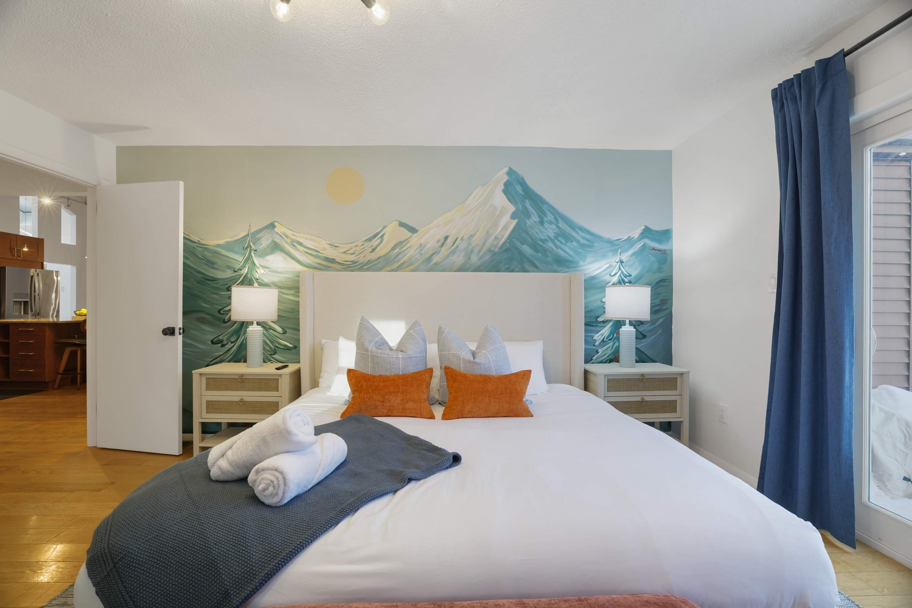 A hotel room with a bed, two bedside tables, a painting on the wall, and a view of the kitchen.