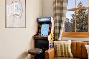 Una habitación con un sofá de cuero marrón, un taburete pequeño, una ventana con cortinas y una máquina de videojuegos.
