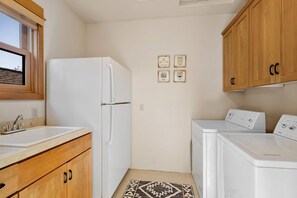 A compact kitchen with a refrigerator, washing machine, and wooden cabinets. - Walk to Downtown With Pool Hot Tub More Access (Sisters)