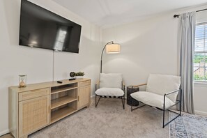 Salón moderno con televisor de pantalla plana, consola de madera, dos sillones blancos, lámpara de pie y vista al exterior a través de una ventana con cortinas.