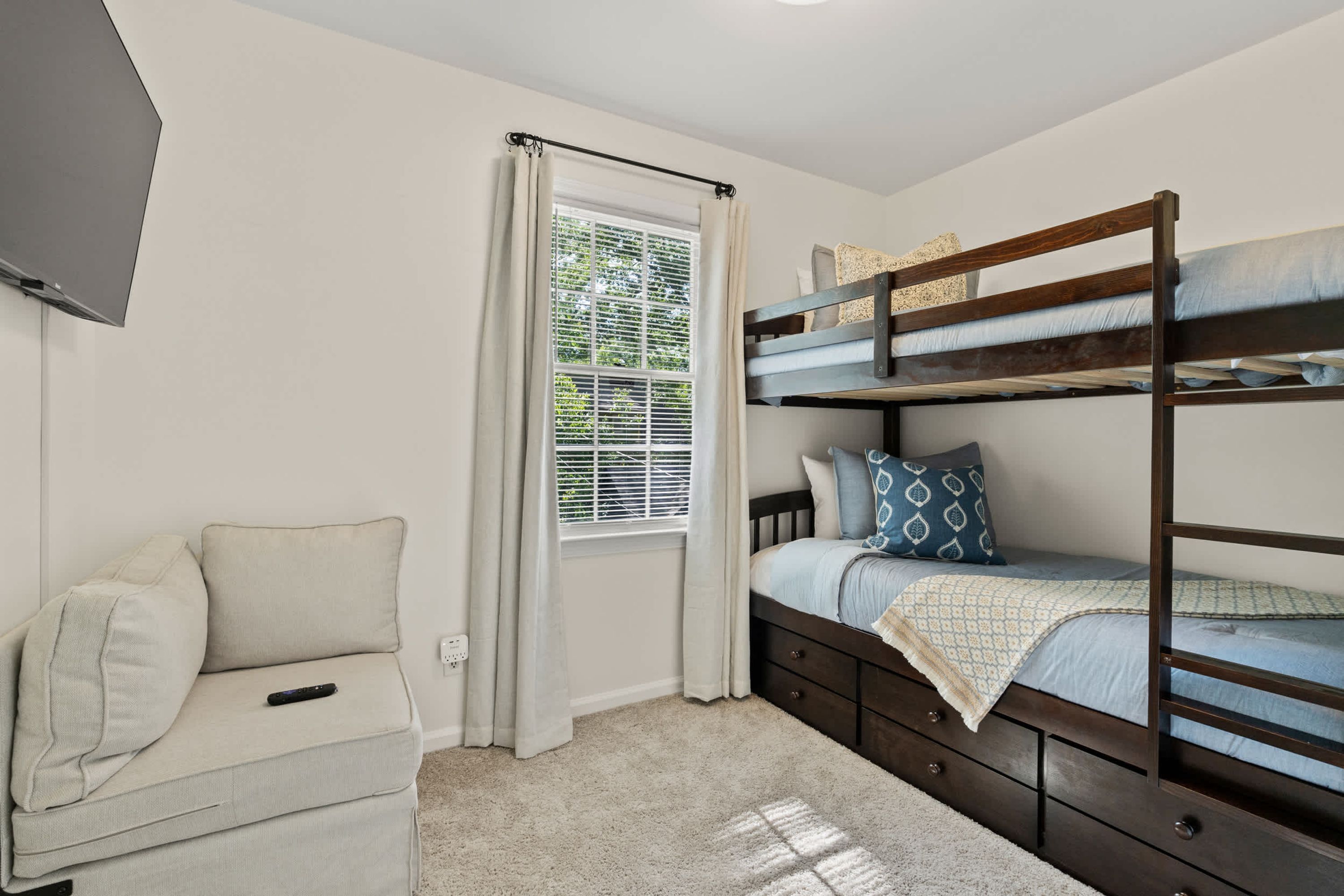 A bedroom with a bunk bed, a sofa, a window with curtains, and a television on the wall.
