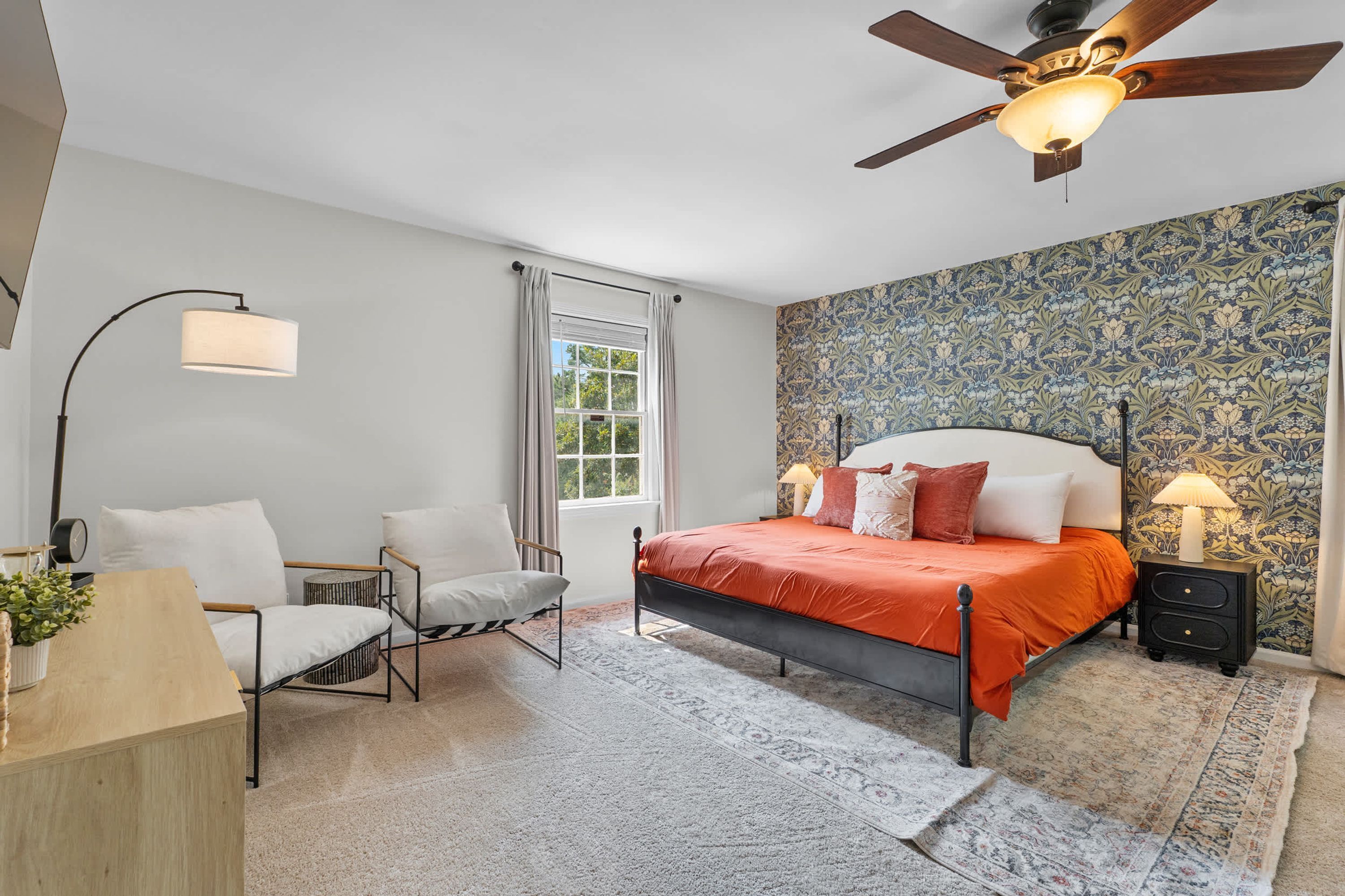 A bedroom with a bed, two chairs, a desk, a nightstand, a lamp, a ceiling fan, and a window with curtains.