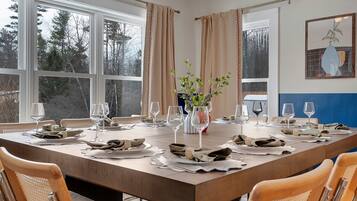 A dining room with a large table set for six, flanked by windows with curtains, a vase with greenery, and a framed picture on the wall.