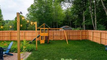 A backyard with a wooden playhouse, a swing set, and a brick patio area.