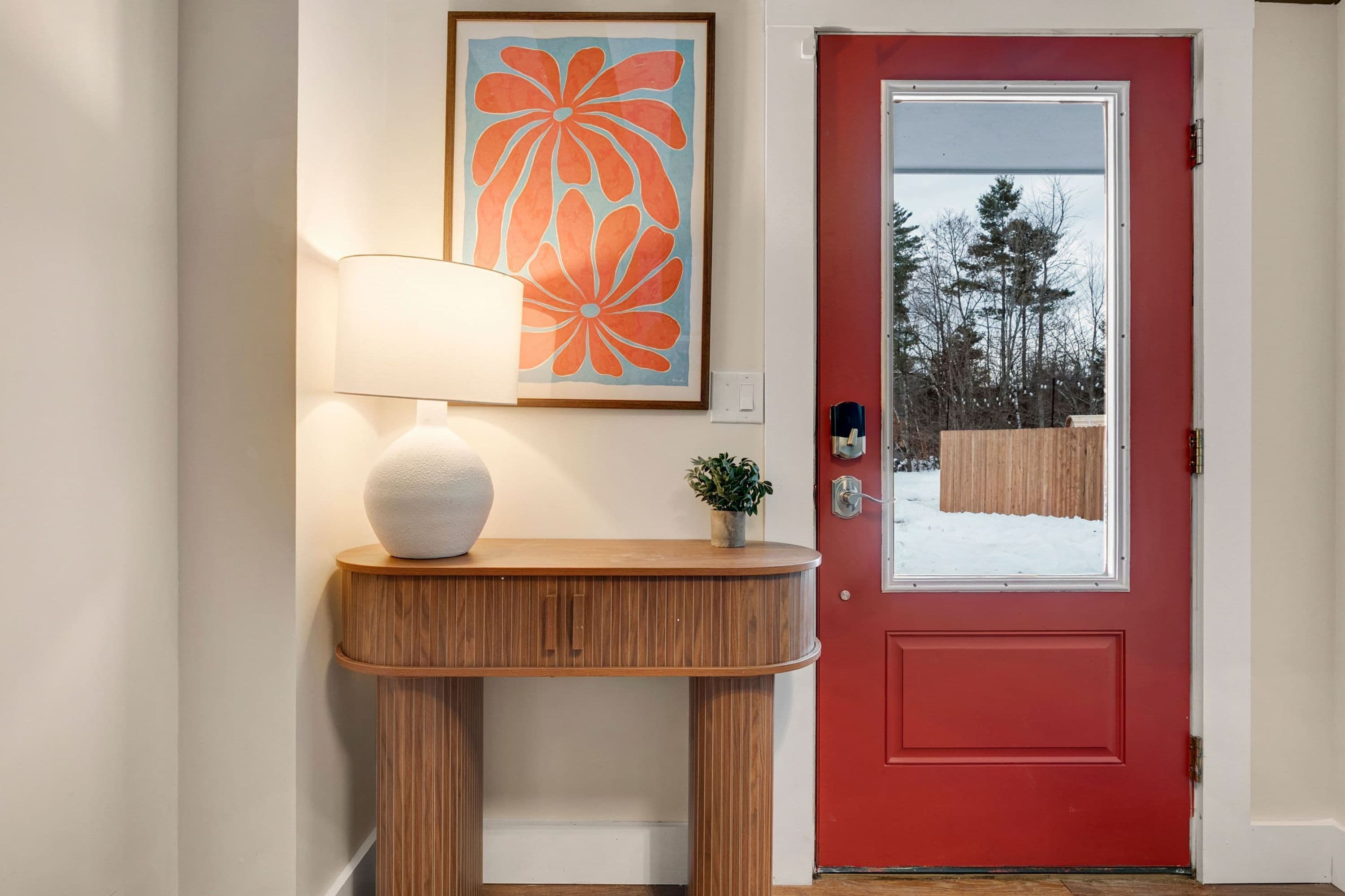 A red door with a glass panel, a wooden console table with a white spherical object, a lamp, and a framed artwork above the console table.