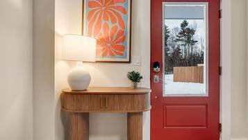 A red door with a glass panel, a wooden console table with a white spherical object, a lamp, and a framed artwork above the console table.