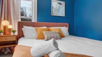 A bedroom with a bed, pillows, a bedside table with lamps, a window, and a painting on the wall.