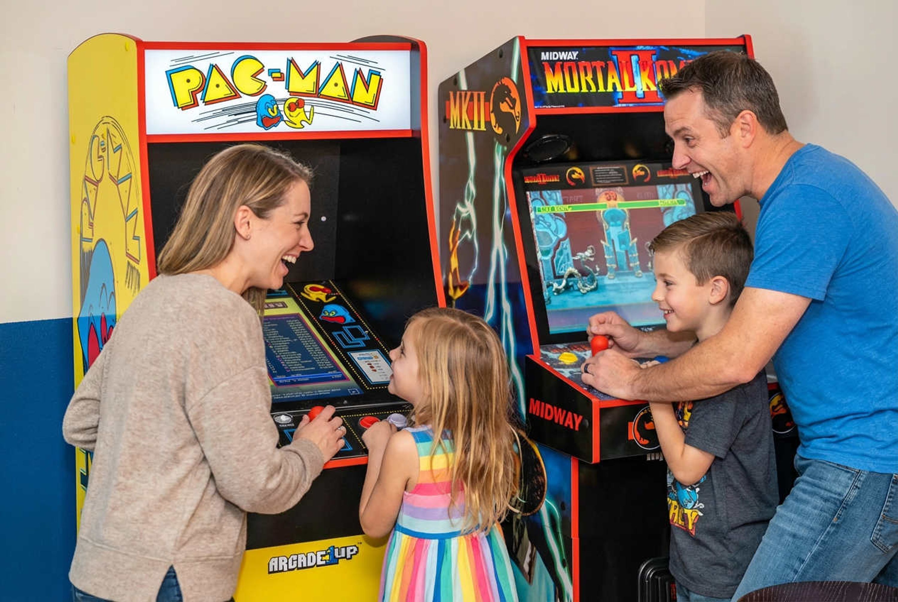 A family playing arcade games, including Pac-Man and Mortal Kombat, with a colorful striped dress and a blue t-shirt.