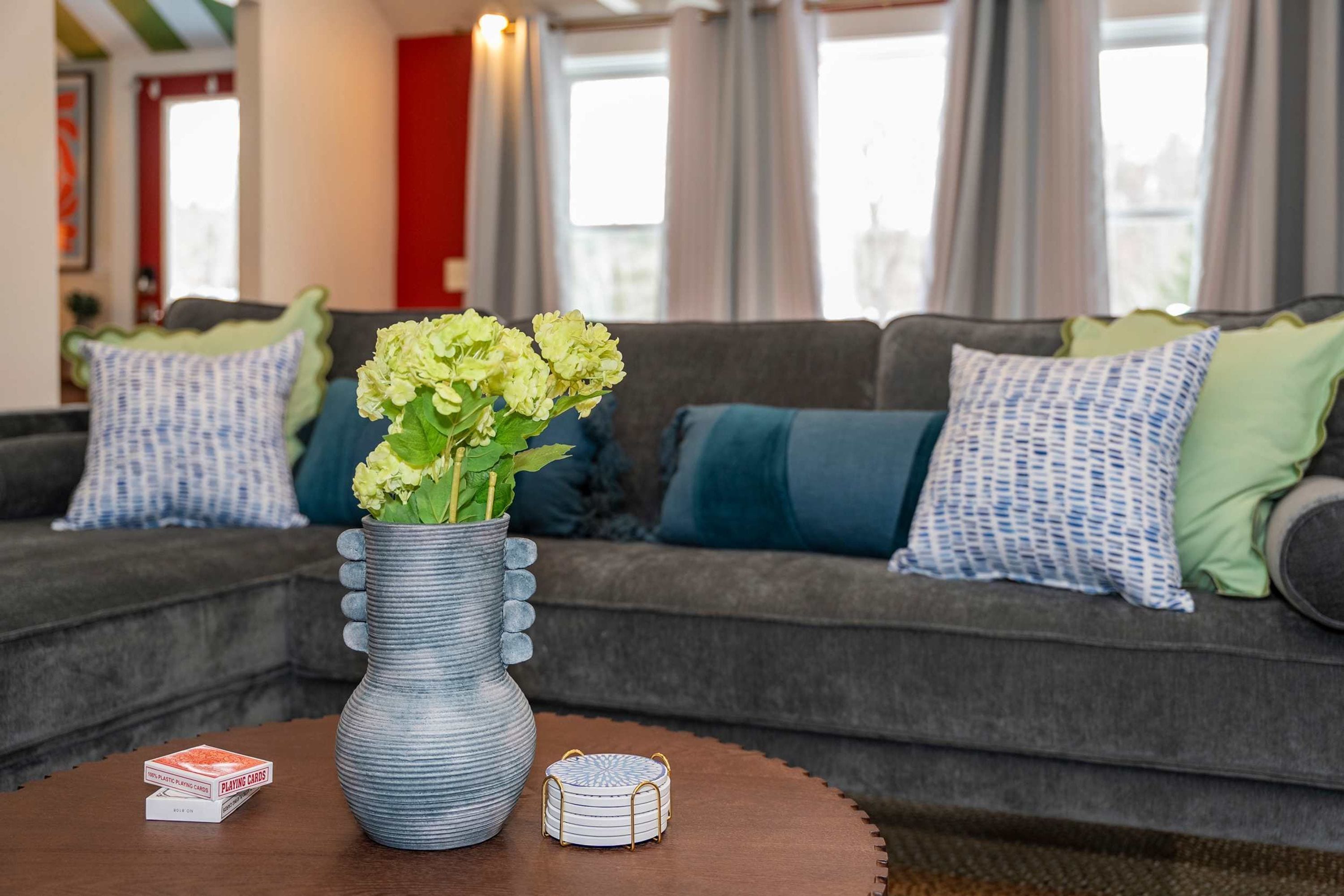 A living room with a grey sofa, green and blue cushions, a vase with flowers, and a wooden table with playing cards and a coaster.