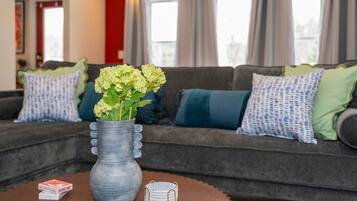 A living room with a grey sofa, green and blue cushions, a vase with flowers, and a wooden table with playing cards and a coaster.