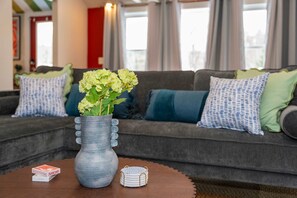 A living room with a grey sofa, green and blue cushions, a vase with flowers, and a wooden table with playing cards and a coaster. - Mountain Getaway Outdoor Games Sauna Hot tub (North Conway)