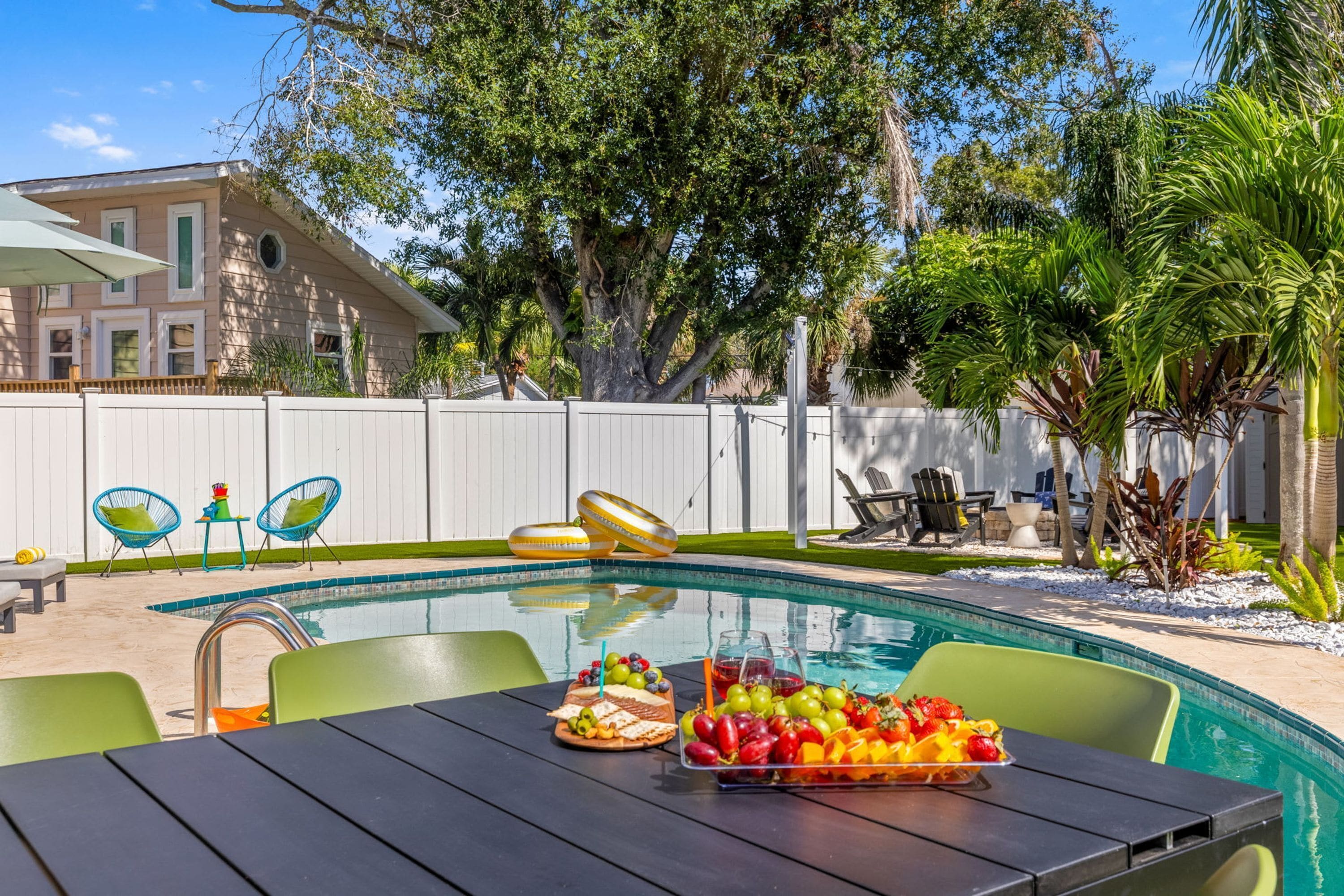 Piscina con mesa dispuesta para una comida, rodeada de mobiliario de exterior y una valla blanca.