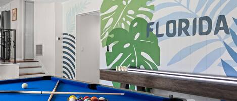 A pool table with balls and a cue stick, a staircase, and a wall with a Florida-themed mural.