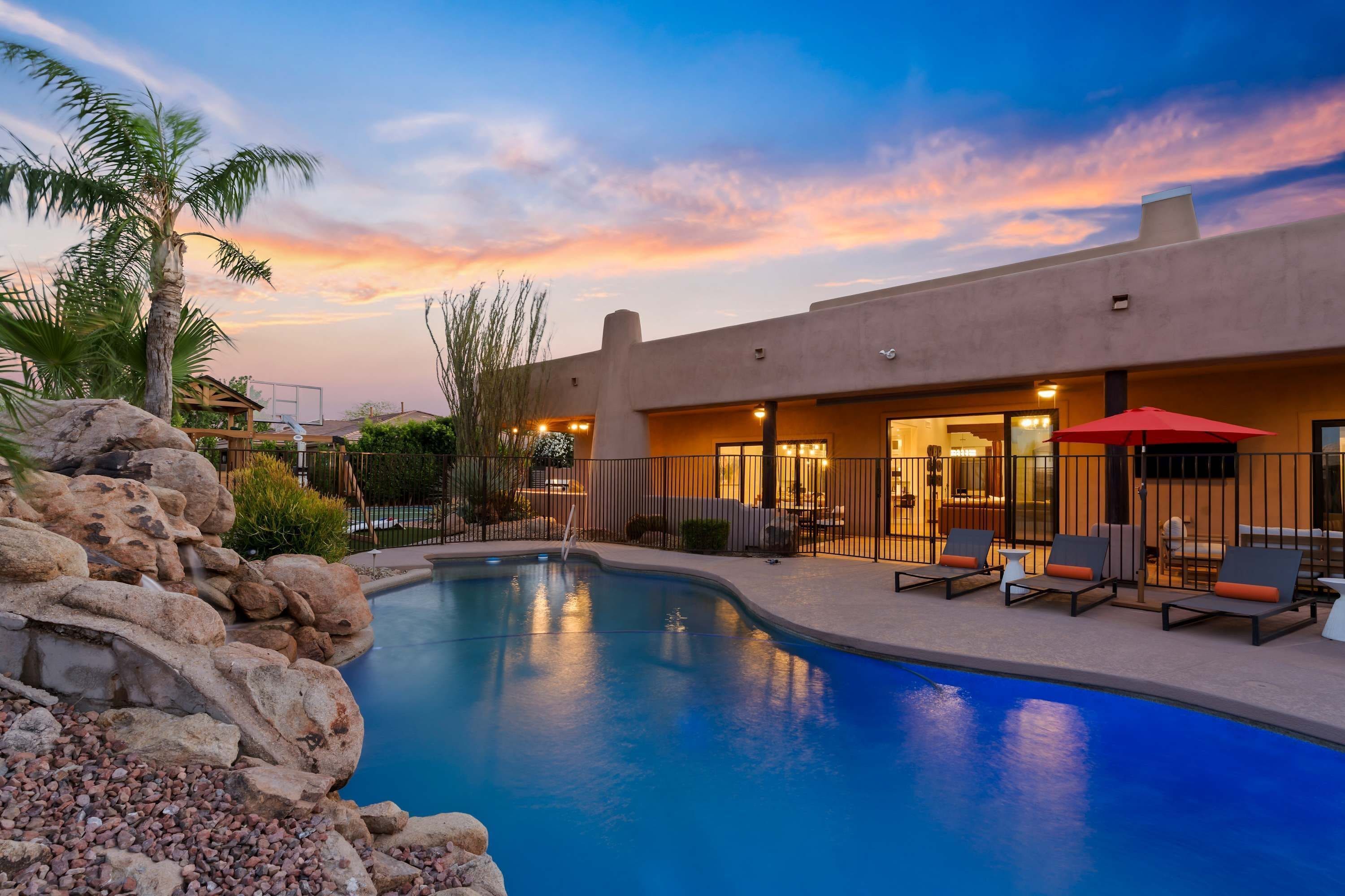 A swimming pool with a stone waterfall feature, surrounded by a patio with lounge chairs and a red umbrella.