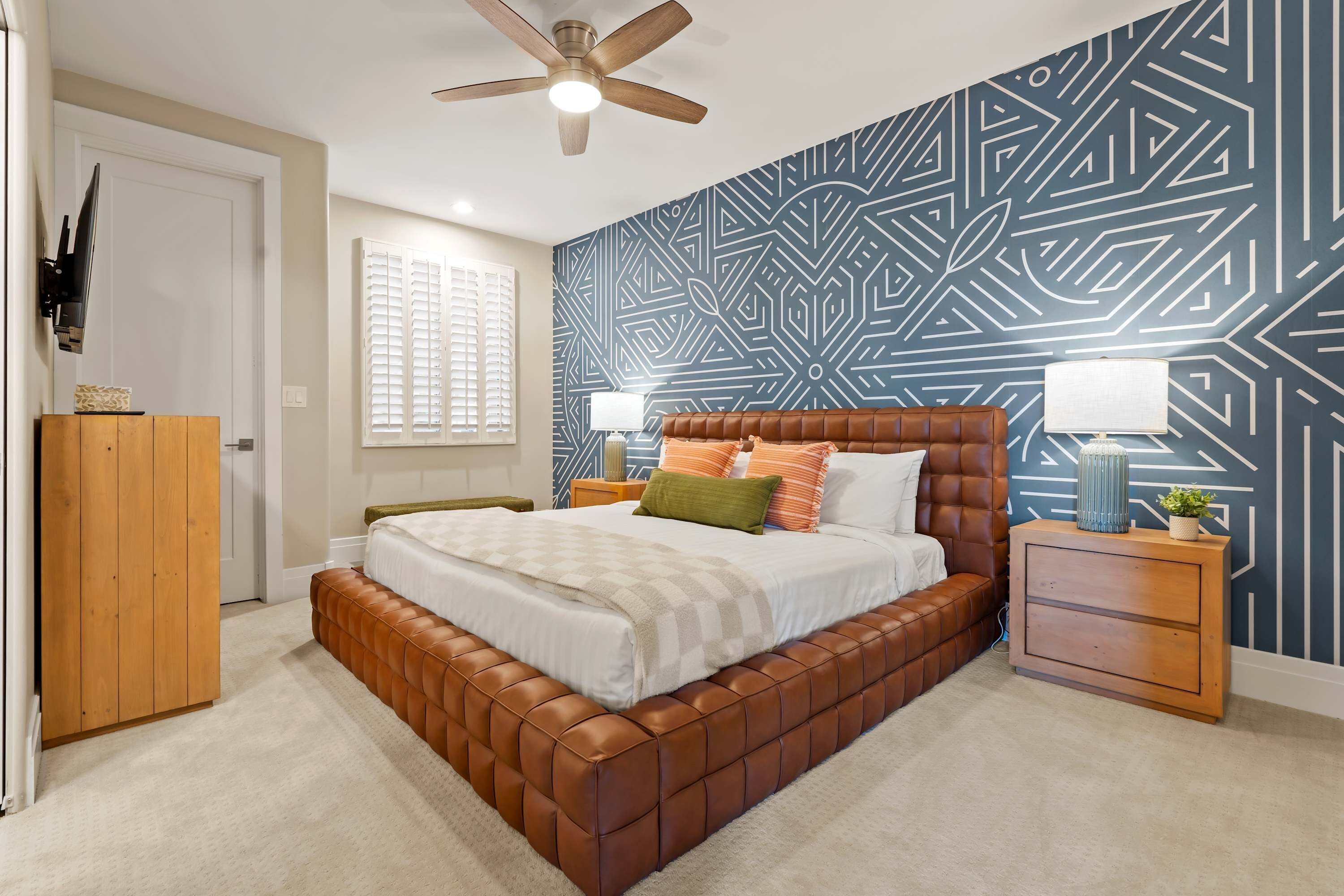 A bedroom with a brown leather bed, a geometric patterned wall, a ceiling fan, and a nightstand with a lamp.