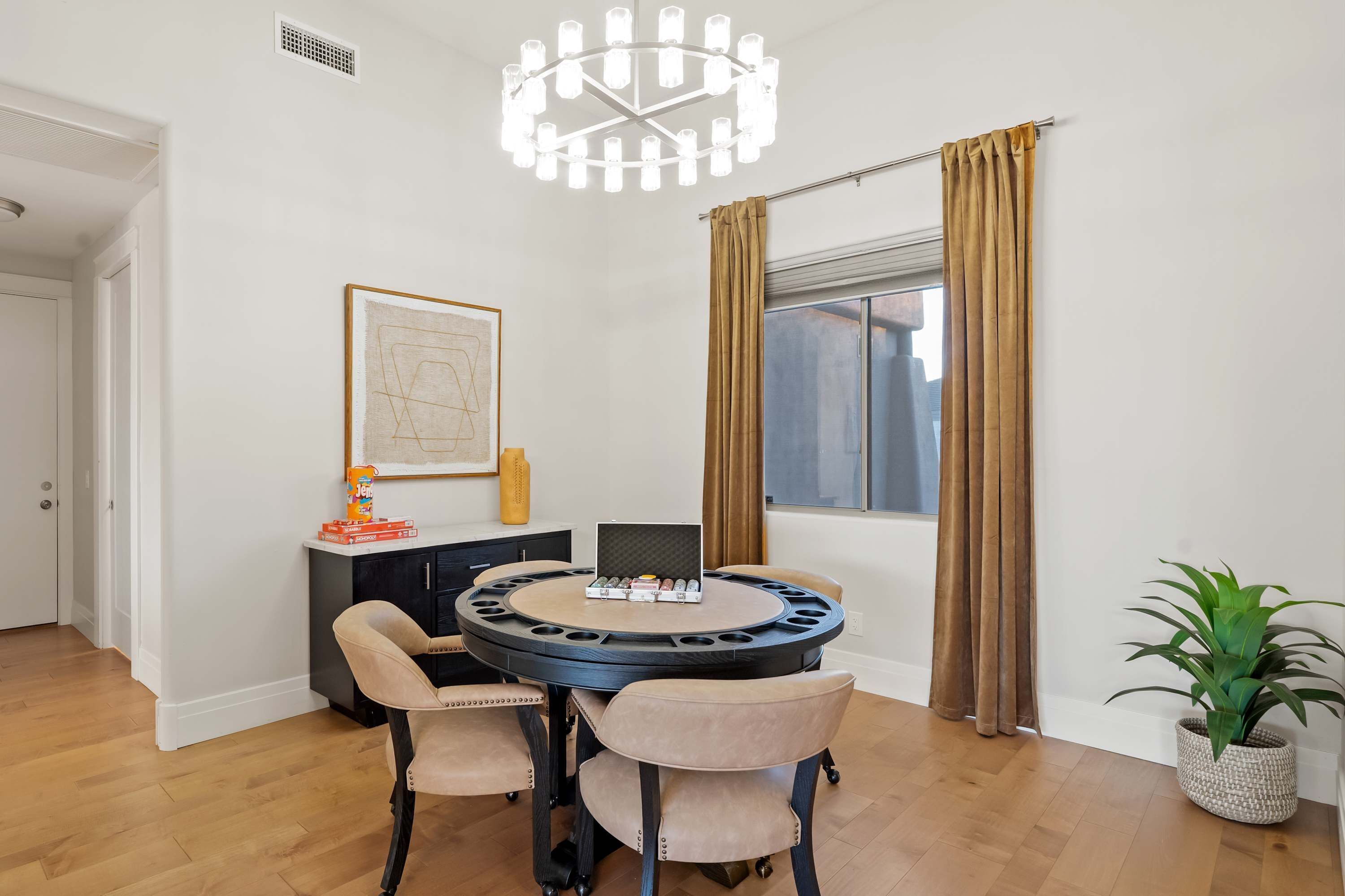 A dining area with a round table, four chairs, a chandelier, a framed picture, a bottle, and a window with curtains.