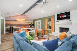 A living room with a fireplace, a mounted TV, and a mural on the wall. - Soak Sizzle Score Pool Games More (San Antonio)