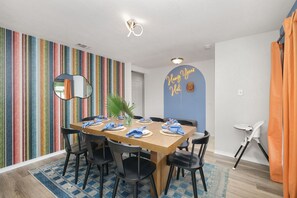 A dining area with a wooden table set for four, surrounded by black chairs. A colorful striped wall and a mirror are visible in the background. - Soak Sizzle Score Pool Games More (San Antonio)