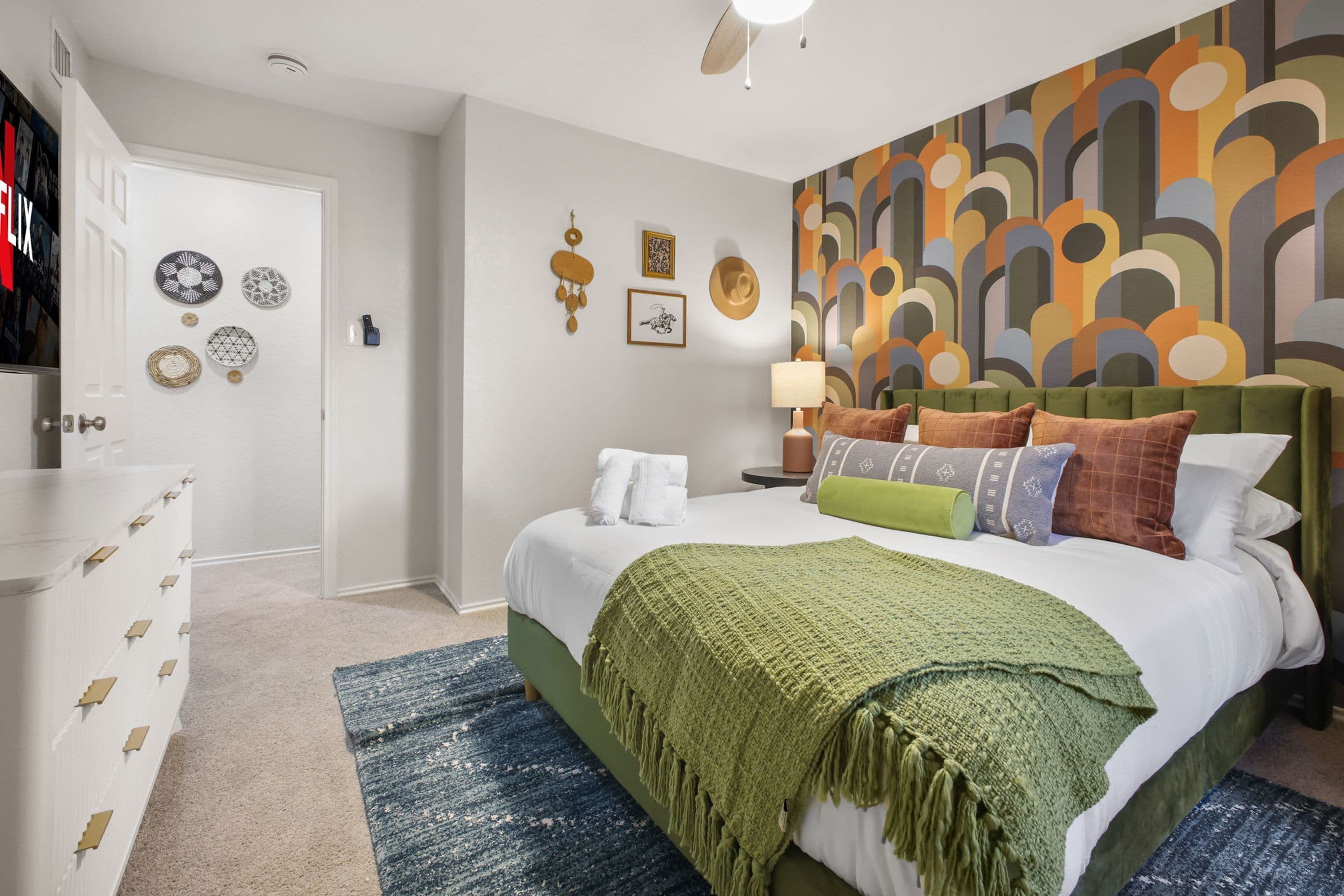 Ein Schlafzimmer mit Bett, Kissen, einer grünen Tagesdecke, einem Kopfteil, einer Wand mit abstrakter Kunst, einem Nachttisch mit Lampe und einer Tür mit Kleiderhaken.