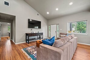 Ein Wohnzimmer mit einem großen Flachbildfernseher an der Wand, einem Ecksofa, einem Holztisch und einem gemusterten Teppich.