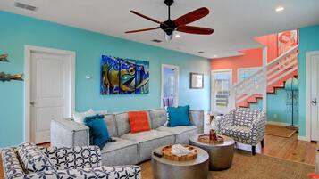 A living room with a grey sofa, patterned armchairs, a ceiling fan, and a colorful abstract painting.