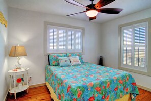 Ein Schlafzimmer mit einem Bett, zwei Fenstern mit Jalousien, einem Deckenventilator, einem Nachttisch mit Lampe und dekorativen Kissen.