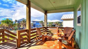A covered outdoor area with wooden chairs and a patterned rug, overlooking a residential area with houses and palm trees.