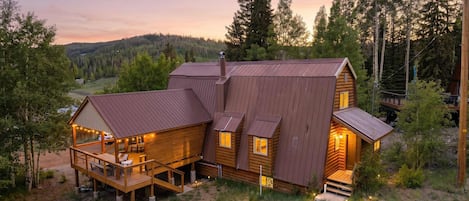 Une cabane dotée d’une terrasse et d’un grand porche, entourée d’arbres et située dans un paysage vallonné.