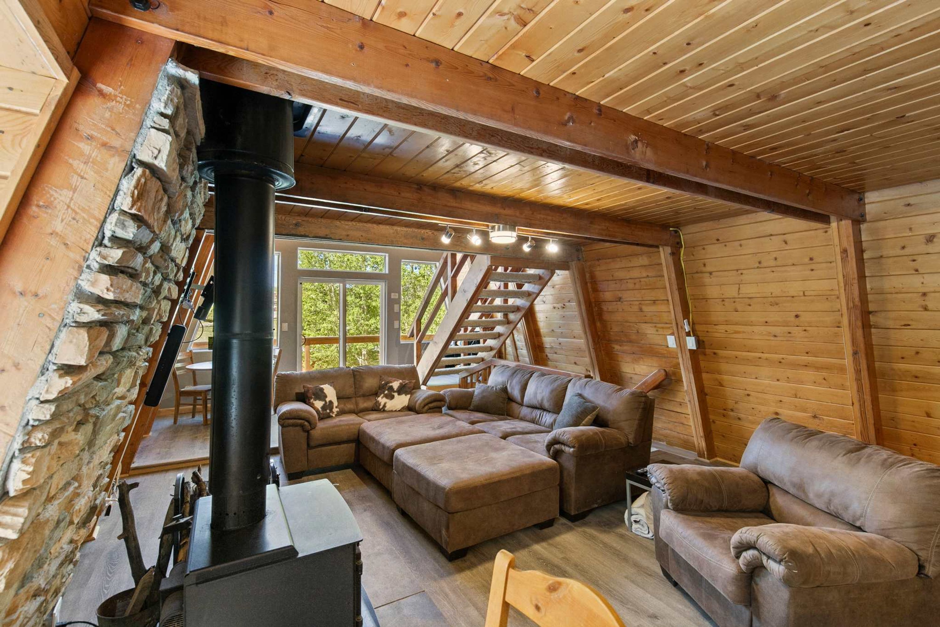 A cozy log cabin interior with a stone wall, a fireplace, a staircase, and a large window.