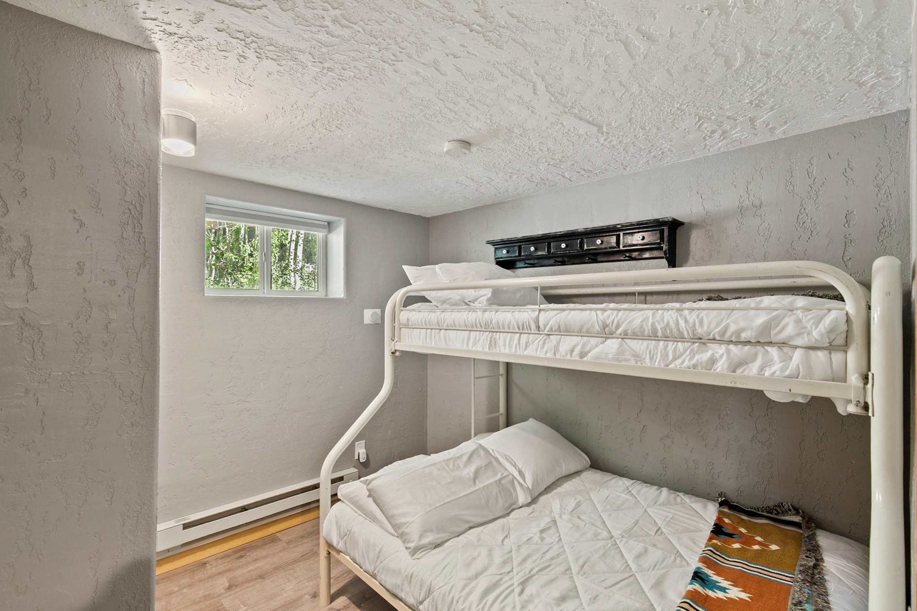 Een slaapkamer met een stapelbed, raam, dressoir en een gebloemd kleed.