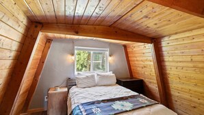 Une chambre avec un plafond en pente, un lit recouvert de linge de lit blanc, une table de chevet, une commode et une fenêtre donnant sur un paysage verdoyant.